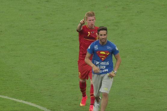 Fußballspieler im roten Trikot zeigt mit dem Finger nach vorn, vor ihm läuft ein Mann im blauen T-Shirt über den Rasen.