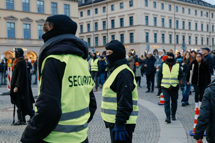 Several individuals wearing masks and safety vests stand in a square in front of a crowd of people and historic buildings in the background.