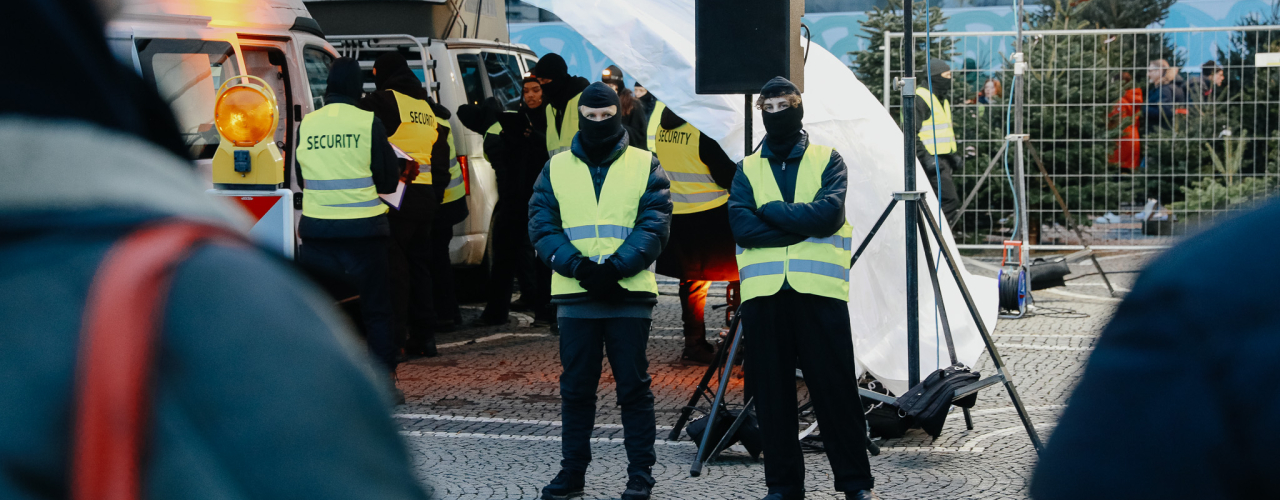 In front of and under a white tent roof, several masked individuals wearing safety vests are standing. Under the tent roof, there are two white vans with their doors open.