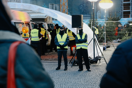 In front of and under a white tent roof, several masked individuals wearing safety vests are standing. Under the tent roof, there are two white vans with their doors open.