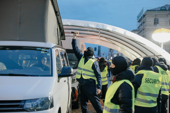 Masked security guards stand next to a white van, and a security officer shines a flashlight into the raised roof of the van.