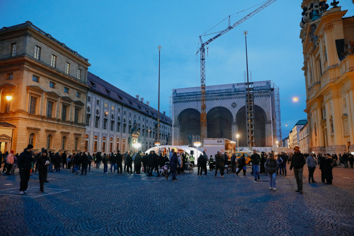 A temporary checkpoint with a tent roof stands in the twilight on a square in front of a historic building. The square is illuminated by four bright lamps. Many people are watching the scene.