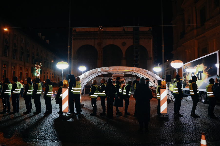 A temporary checkpoint with a tent roof is located on a square in front of a historic building. A row of security personnel stands in front of the nighttime scene, which is illuminated by four bright lamps.