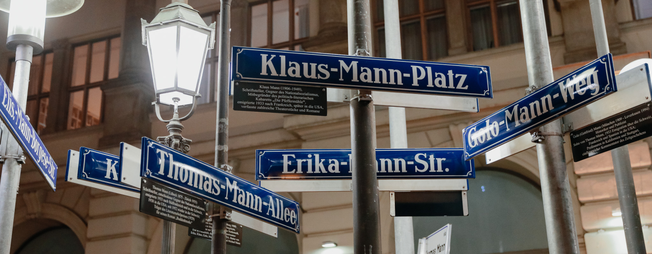 Blick auf einige Straßenschilder mit Namen der Familie Mann an unterschiedlichen Straßenlaternen befestigt. Im Hintergrund eine historische Häuserfassade.
