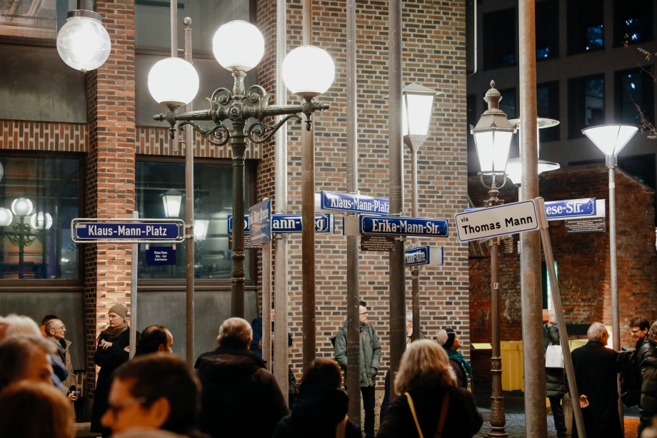 Einige Menschen stehen zwischen unterschiedlichen leuchtenden, teils historischen Straßenlaternen vor einer Backsteinfassade. An den Straßenlaternen sind Straßenschilder mit verschiedenen Namen der Familie Mann befestigt.