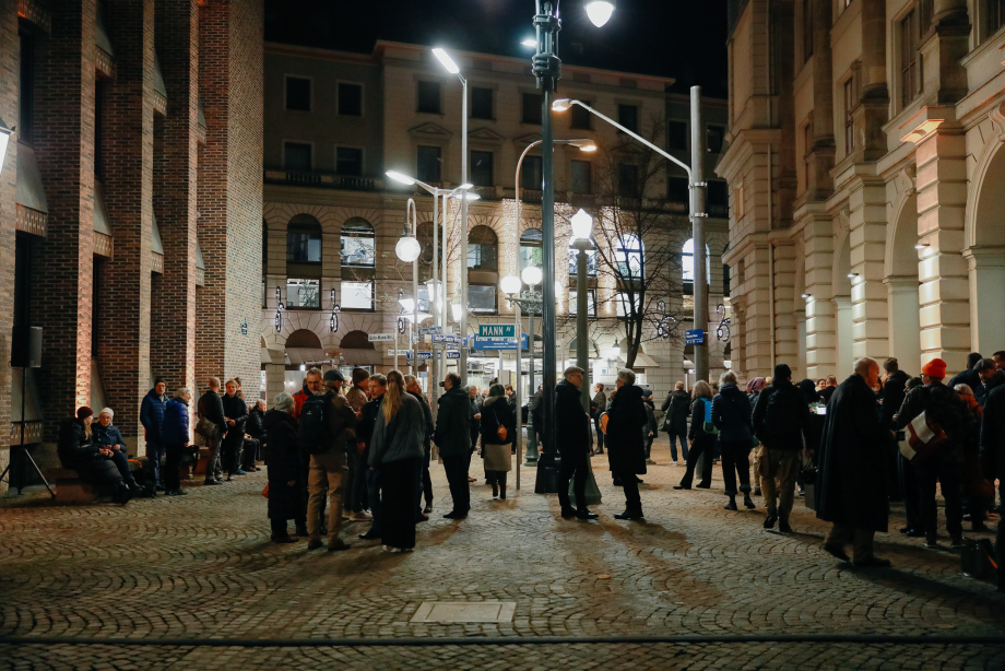 Viele Menschen stehen zwischen historischen Häuserfassaden auf einem Platz, der von einigen unterschiedlichen Straßenlaternen beleuchtet ist. An diesen sind verschiedene Straßenschilder mit Namen der Familie Mann befestigt.