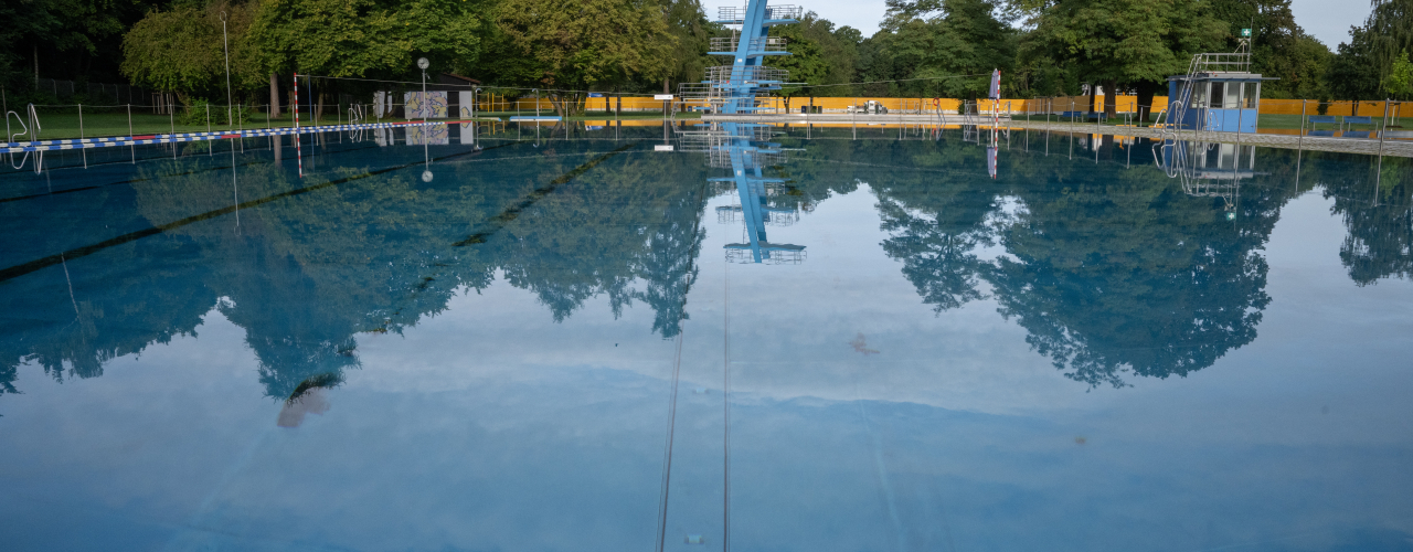 Leeres Freibad mit Sprungturm, der sich im ruhigen Wasser spiegelt, umgeben von Bäumen.