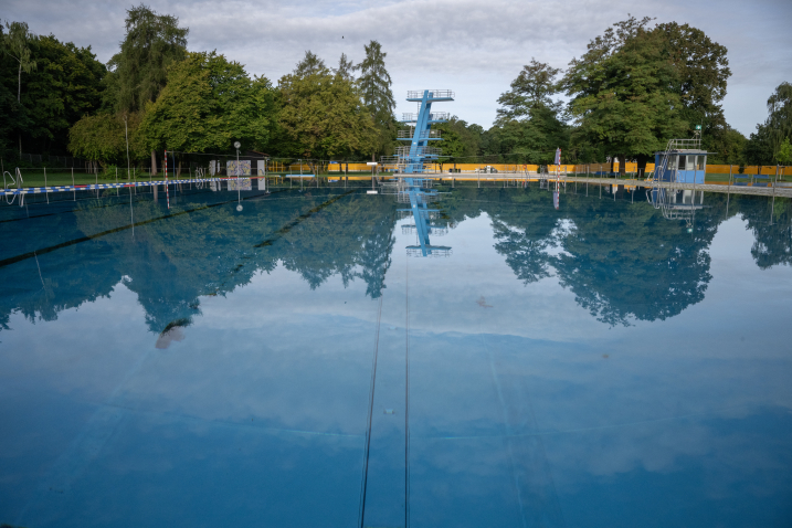 Leeres Freibad mit Sprungturm, der sich im ruhigen Wasser spiegelt, umgeben von Bäumen.