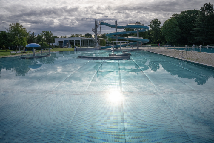 Leeres Becken im Freibad mit geschlängelter Rutsche, im Hintergrund Bäume und Wolkenhimmel.