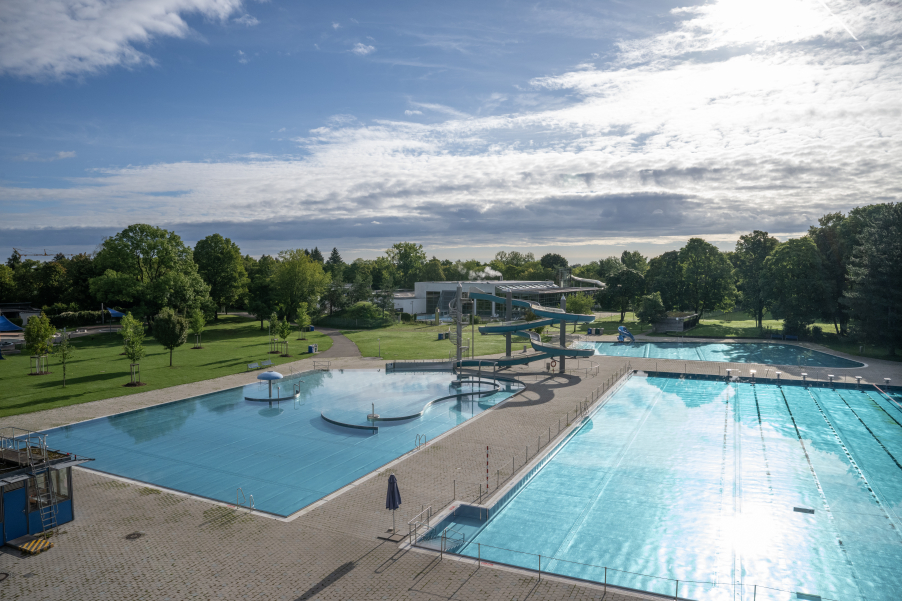 Leeres Freibad mit drei Becken und langer, geschlängelter Rutsche, umgeben von Bäumen.