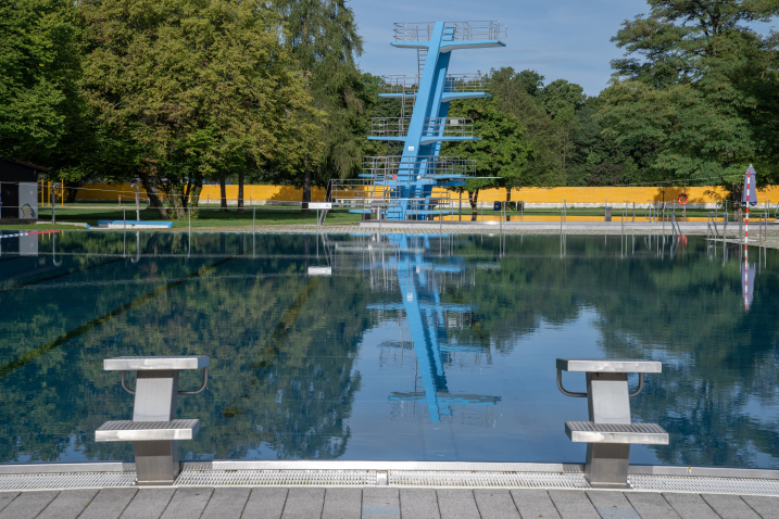 Leeres Freibad mit Sprungturm, der sich im ruhigen Wasser spiegelt, umgeben von Bäumen.