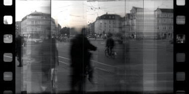 Black-and-white photo on a film strip: Blurry figures and a cyclist in a bustling city square surrounded by old buildings.