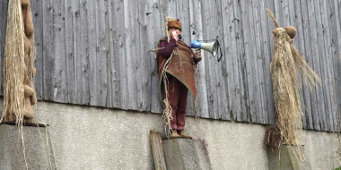 Four costumed figures made of straw and natural materials stand on concrete pillars in front of a wooden barn; one of them is a real person holding a megaphone.