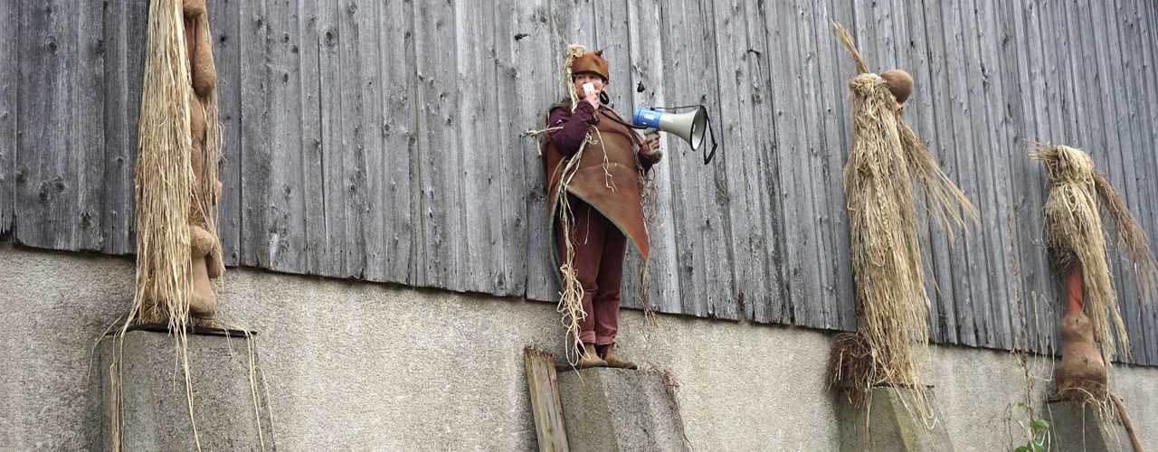 Four costumed figures made of straw and natural materials stand on concrete pillars in front of a wooden barn; one of them is a real person holding a megaphone.