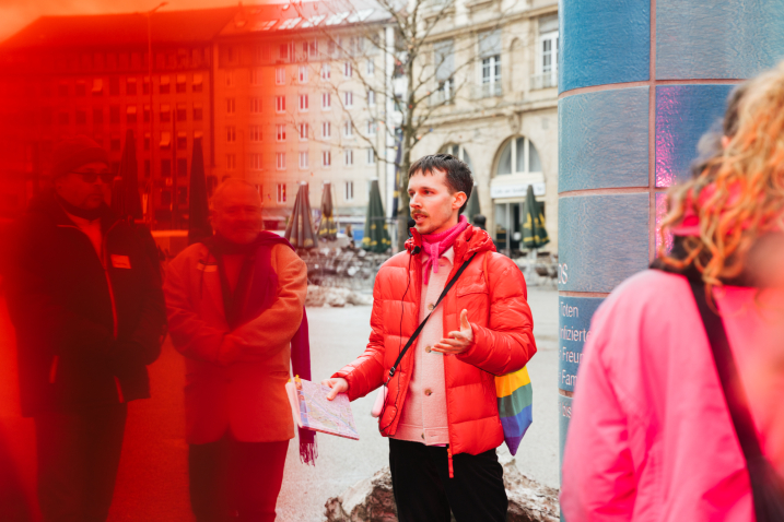 Eine Person in roter Daunenjacke mit einem Stofftasche in Regenbogenfarben steht vor einer gekachelten Säule auf einem Stadtplatz und spricht zu einer Gruppe von Menschen.