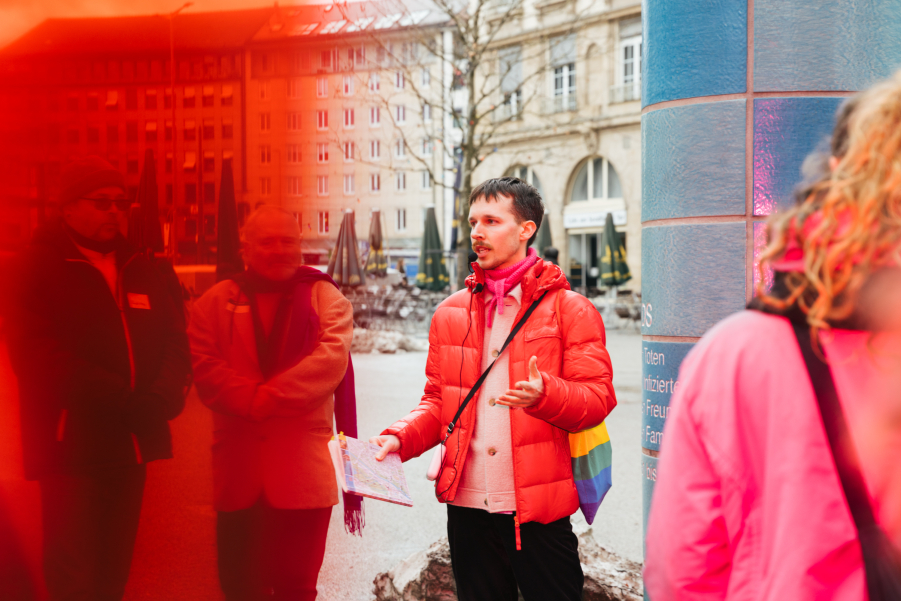 Eine Person in roter Daunenjacke mit einem Stofftasche in Regenbogenfarben steht vor einer gekachelten Säule auf einem Stadtplatz und spricht zu einer Gruppe von Menschen.