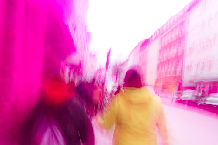 Ein unscharfes Bild zeigt Personen von hinten, die auf dem Gehweg spazieren. Links und rechts sind Gebäudefassaden. Das Bild ist rot-transparent überlagert.