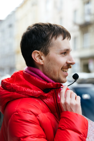 Der Gay Guide Anian Halder ist im Profil zu sehen. Er trägt eine rote Jacke und einen rosa Schal. Mit seiner Hand berührt er ein Mikrofon, das an seiner Jacke angebracht ist.