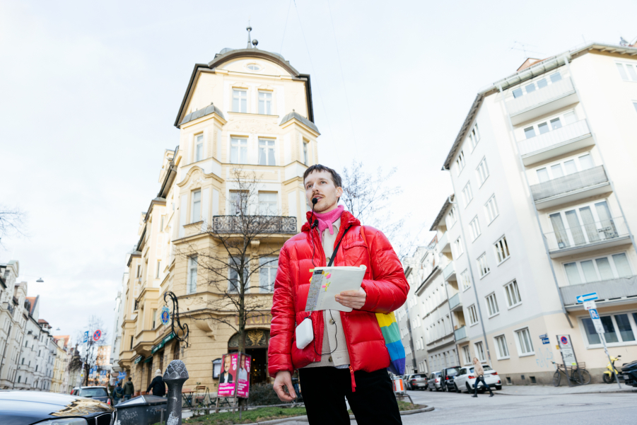 In der Mitte des Bildes ist der Gay Guide Anian Halder. Er steht vor einem gelben Gebäude. In der Hand hält er ein Heft.