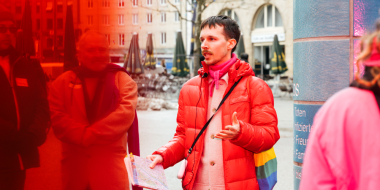 A person wearing a red down jacket and carrying a rainbow-colored bag stands in front of a tiled column in a city square and speaks to a group of people.