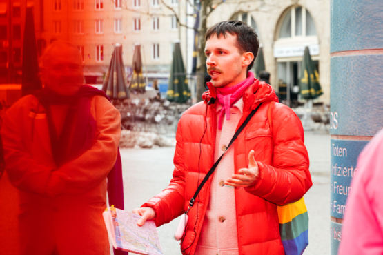 Eine Person in roter Daunenjacke mit einem Stofftasche in Regenbogenfarben steht vor einer gekachelten Säule auf einem Stadtplatz und spricht zu einer Gruppe von Menschen.