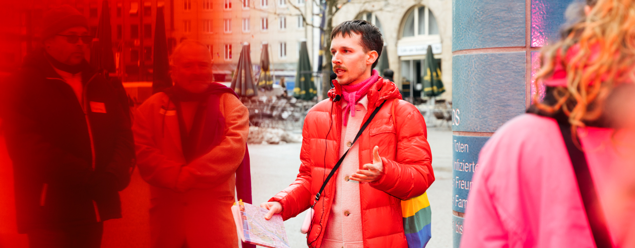 Eine Person in roter Daunenjacke mit einem Stofftasche in Regenbogenfarben steht vor einer gekachelten Säule auf einem Stadtplatz und spricht zu einer Gruppe von Menschen.