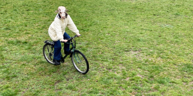 A person wearing a sheep mask is riding a bicycle across a meadow.